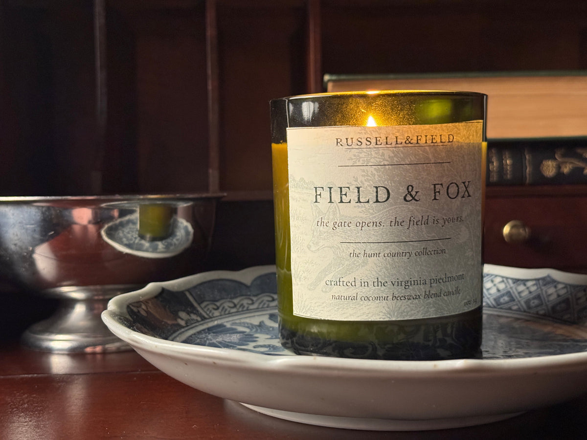 A Russell & Field 'Field & Fox' scented candle in a dark amber glass jar, lit and resting on a white ceramic plate with blue patterns on a wooden table, with a dark wood bookshelf in the background.