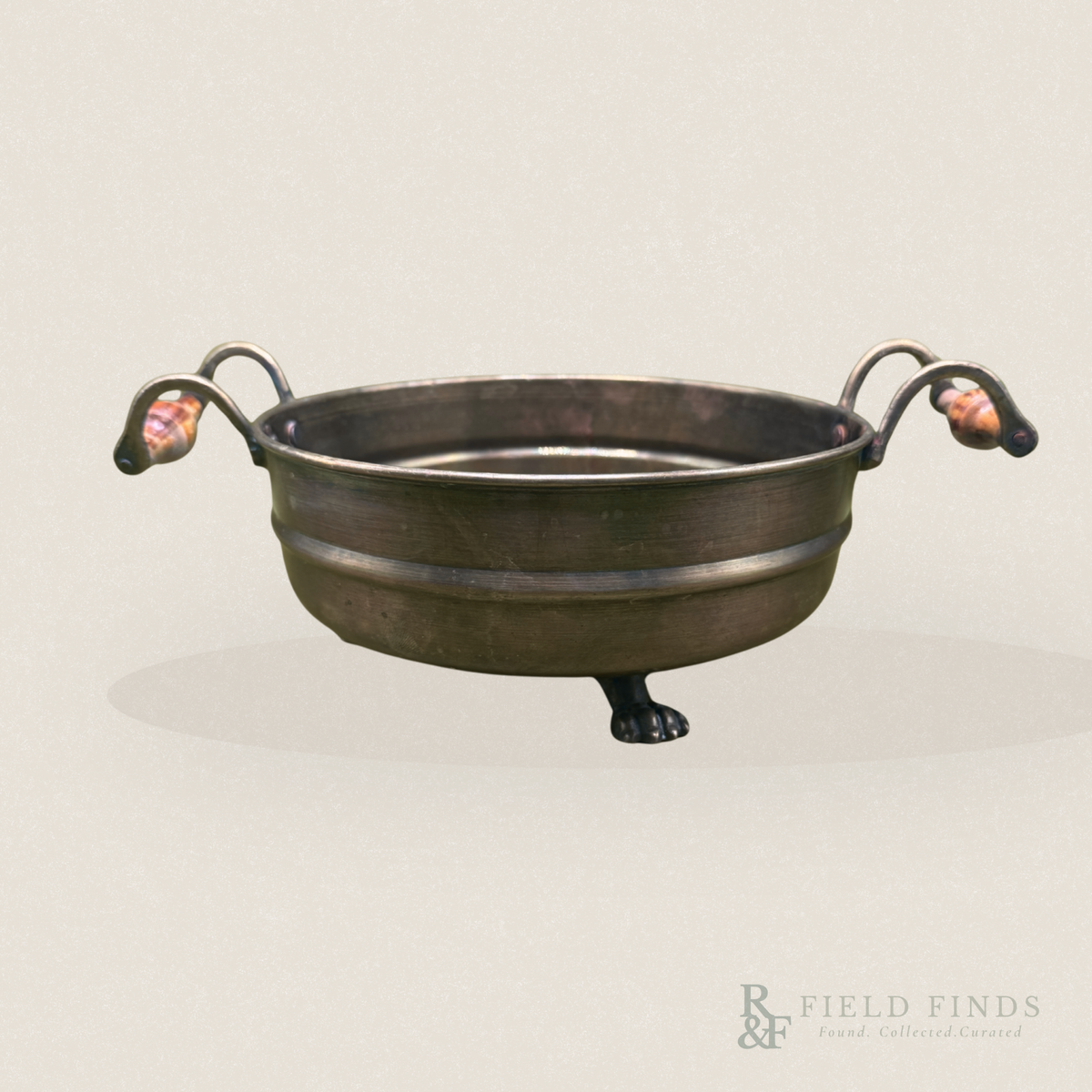 A small dark brass bowl with two looped side handles and decorative claw-like feet, shown on a plain light beige background. A Field Finds watermark appears in the lower right corner.