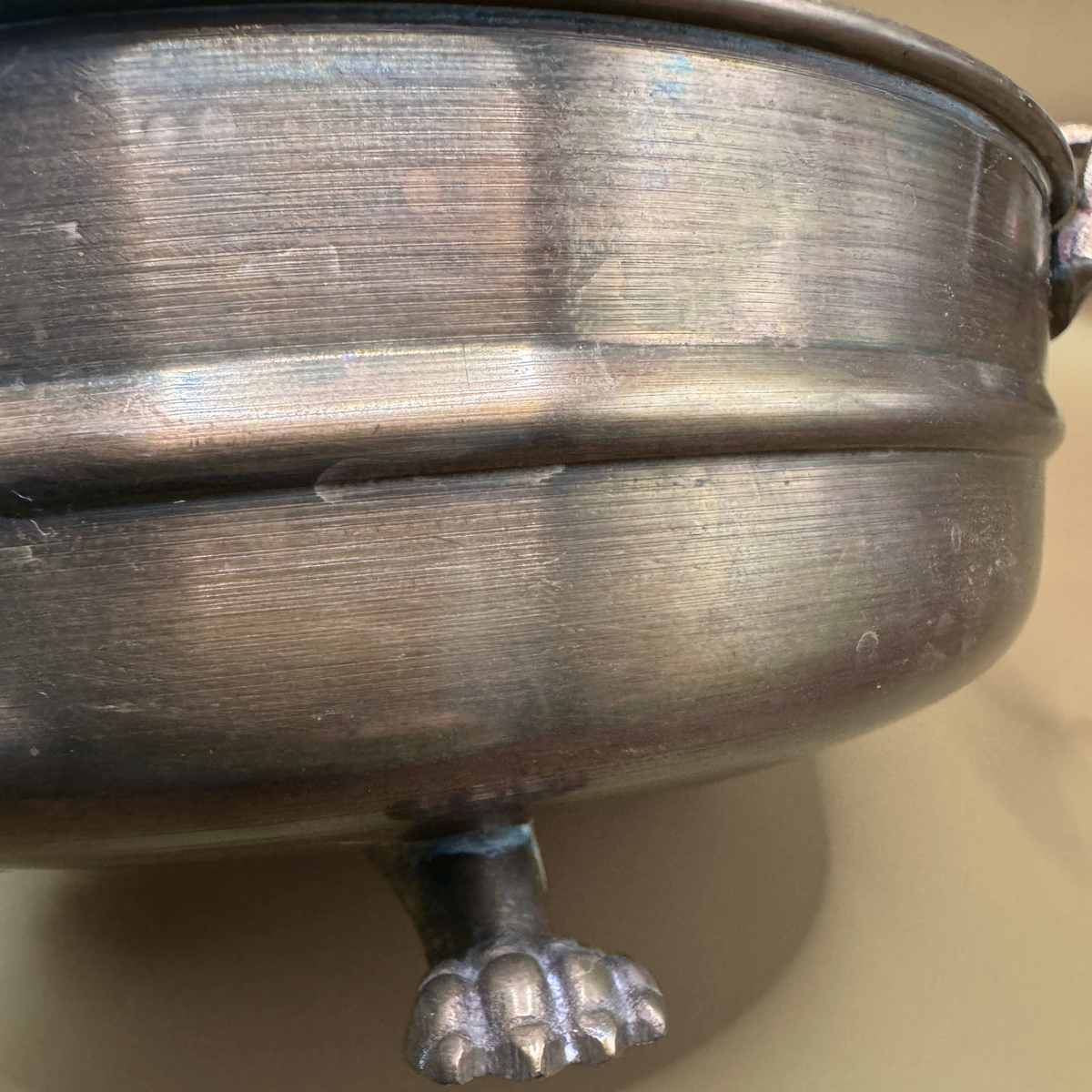 Close-up of a brass footed  cachepot with a paw-shaped foot, set against a plain beige background.