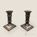 Column Candlesticks-Pair. A pair of vintage dark metal candlestick holders with square bases, shown on a light beige background with a Field Finds watermark in the lower right corner.