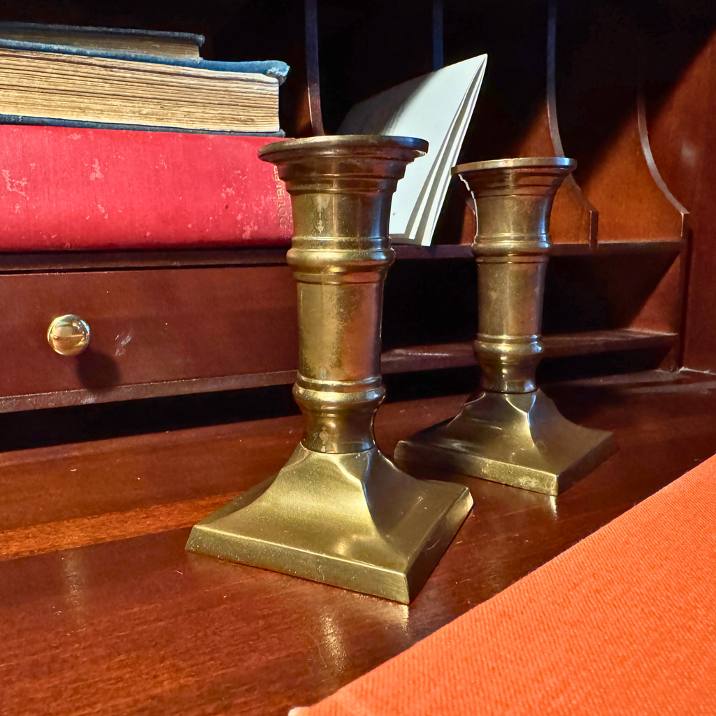 Column Candlesticks-Pair. Two vintage brass candlesticks with a square flared base and ribbed stem, displayed on a polished dark wood desk next to books.