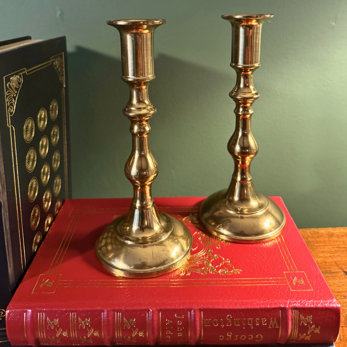 Georgian Style brass candlesticks displayed on top of a red vintage book, with a dark green wall in the background.