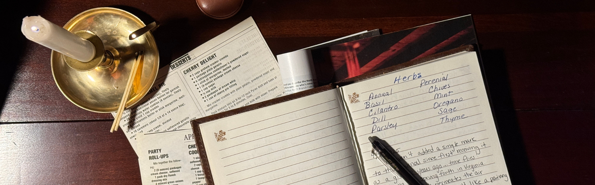 An open journal on a dark wooden table, with handwritten notes  and a black pen resting on it. A brass candle holder with a lit white candle and printed recipe sheets, arranged around it in a warm, rustic setting.