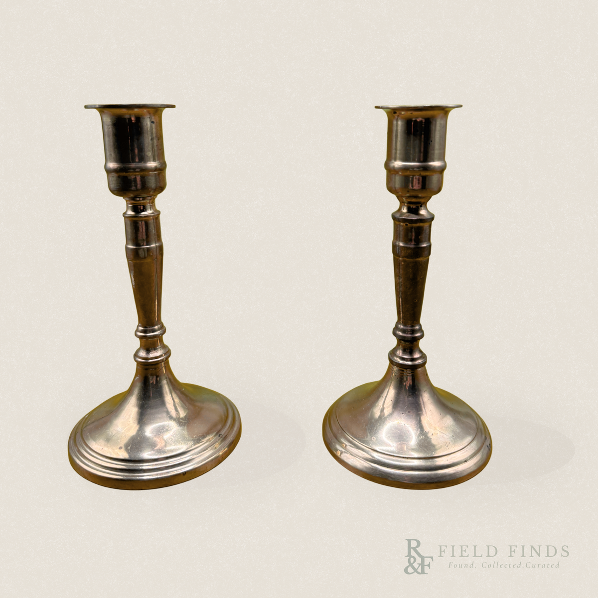 Tapered Brass Candlesticks-Pair. A pair of vintage brass metal candlesticks with flared circular bases and turned decorative stems, shown on a plain light beige background. A 'Field Finds' watermark is visible in the bottom right corner.
