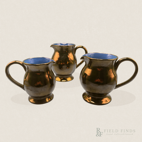 Three vintage Creigiau Pottery Welsh copper lustreware jugs in graduated sizes, featuring reflective bronze-copper finishes, cobalt blue interiors, curved handles, and pouring spouts, arranged against a warm ivory background.